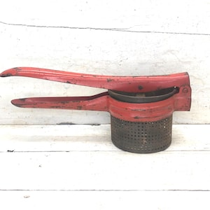May include: A vintage red potato ricer with a perforated base and long handles. The kitchen tool is made of metal and shows signs of wear, suggesting it's an antique. The ricer is designed for mashing potatoes and other soft foods.