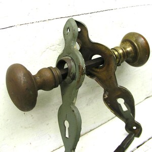 May include: A vintage door hardware set featuring two round brass knobs connected by a square metal spindle. The assembly includes two decorative metal backplates, one finished in a muted sage green paint and the other in aged brass, each with a keyhole.