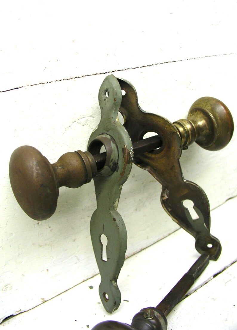 May include: A vintage door hardware set featuring two round brass knobs connected by a square metal spindle. The assembly includes two decorative metal backplates, one finished in a muted sage green paint and the other in aged brass, each with a keyhole.