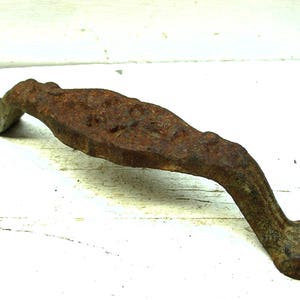 May include: A rustic, antique door handle made of heavily rusted metal. The handle has a curved, elongated shape with decorative detailing and mounting holes on each end. The color is a deep brown with areas of lighter rust.