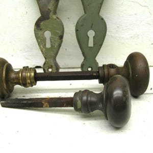 May include: Two vintage metal door knobs with dark brown finishes and attached square spindles, accompanied by two matching sage green metal backplates featuring keyhole cutouts. The hardware displays signs of age and wear on the surfaces.