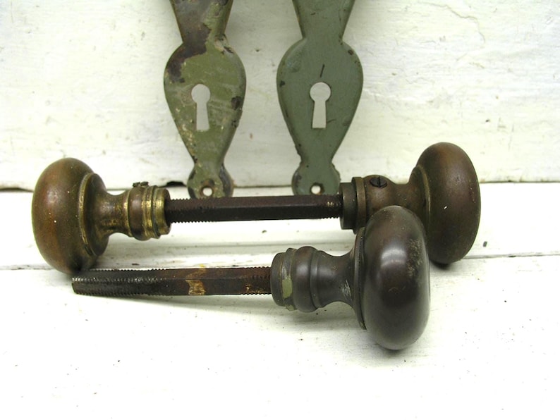 May include: Two vintage metal door knobs with dark brown finishes and attached square spindles, accompanied by two matching sage green metal backplates featuring keyhole cutouts. The hardware displays signs of age and wear on the surfaces.