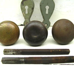 May include: Three round metal door knobs with varying patinas, including one brass-toned and two dark brown finishes. Two matching metal keyhole escutcheon plates with a light green finish and two rectangular metal spindles are included in the set.