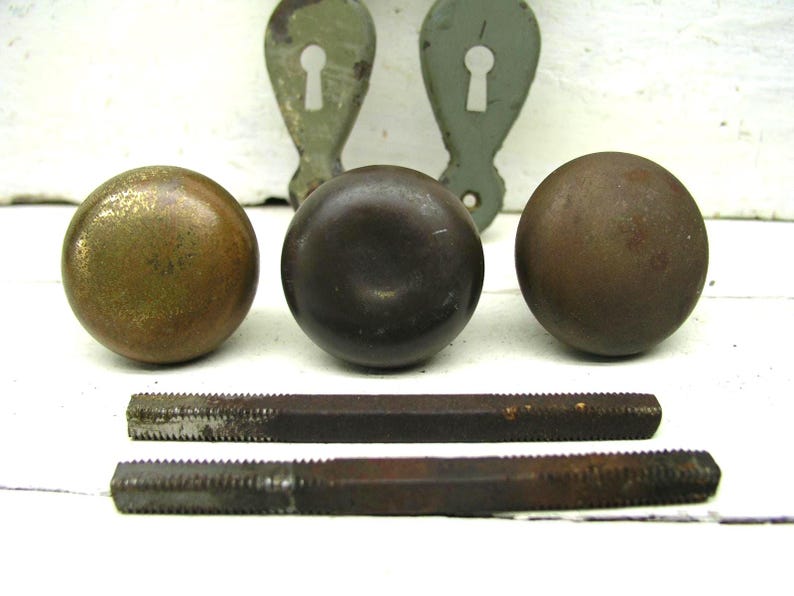 May include: Three round metal door knobs with varying patinas, including one brass-toned and two dark brown finishes. Two matching metal keyhole escutcheon plates with a light green finish and two rectangular metal spindles are included in the set.