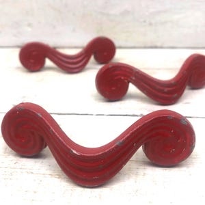 May include: Four vintage red drawer pulls with a wavy, scroll-like design. The pulls have a distressed finish, showing signs of wear and age. These decorative hardware pieces are ideal for adding a touch of retro charm to furniture.