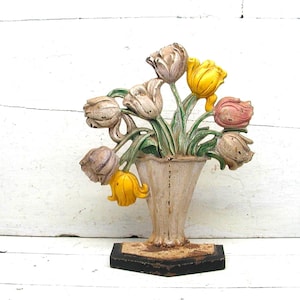 May include: A cast metal doorstop featuring a bouquet of tulips in a vase. The tulips are painted in shades of yellow, pink, and muted white, with green stems and leaves. The vase is off-white, and the base is a solid, dark, rectangular block.