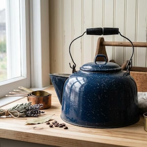 May include: A dark blue speckled enamel tea kettle with a black handle, accompanied by a set of copper measuring cups, a bundle of dried lavender, a single bay leaf, a small brass cup, and scattered coffee beans on a wooden surface.