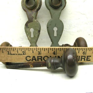 May include: Two sage green metal door escutcheons with keyholes and two dark brown metal door knobs with spindles. A wooden ruler is positioned in front of the hardware, showing the knobs are approximately 2.25 inches in diameter.