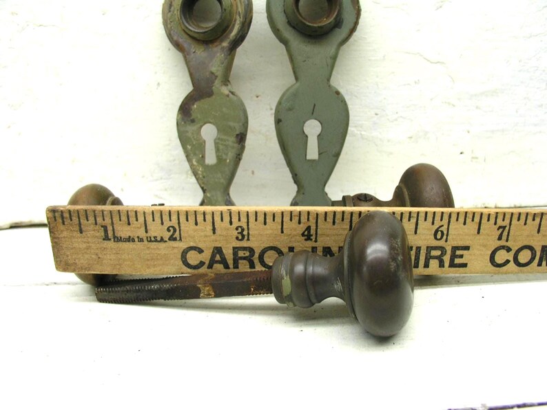 May include: Two sage green metal door escutcheons with keyholes and two dark brown metal door knobs with spindles. A wooden ruler is positioned in front of the hardware, showing the knobs are approximately 2.25 inches in diameter.