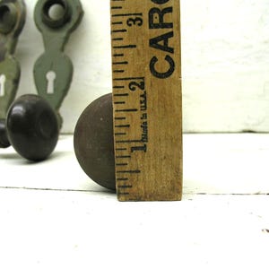May include: A dark brown round door knob positioned next to a wooden ruler showing a diameter of approximately 2.25 inches. In the background, two sage green metal door plates with keyholes are visible, each featuring a circular top opening.