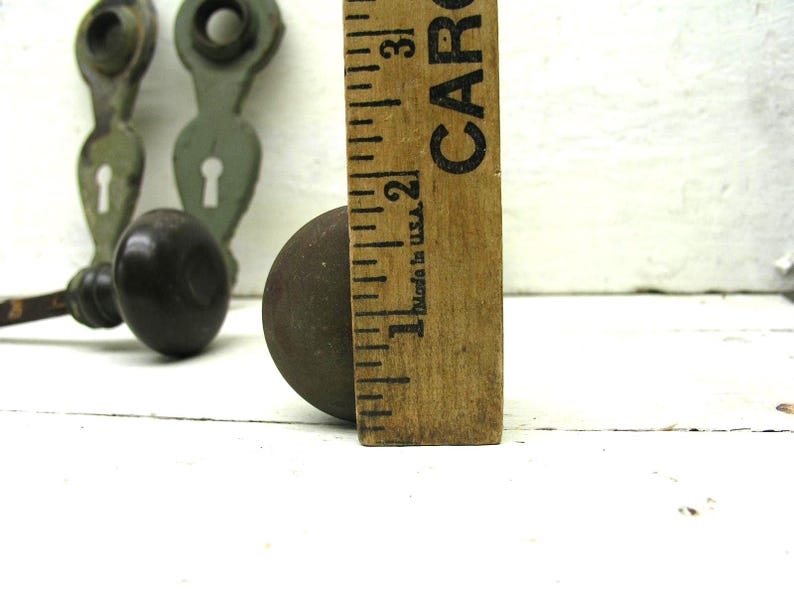 May include: A dark brown round door knob positioned next to a wooden ruler showing a diameter of approximately 2.25 inches. In the background, two sage green metal door plates with keyholes are visible, each featuring a circular top opening.