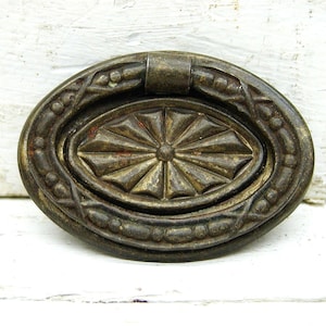 May include: An oval-shaped, antique brass drawer pull with a decorative, starburst-shaped center and a raised, beaded edge.