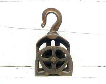 May include: A rusty cast iron pulley with a hook on top. The pulley has a wheel with a grooved rim and a metal frame.