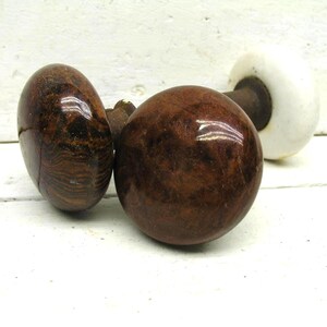 May include: Two round door knobs with a polished dark brown wood grain finish and a metallic spindle connecting them to a single white ceramic base plate. The knobs have a smooth, reflective surface and a spherical shape, showing natural wood patterns.