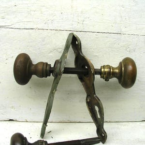 May include: A set of antique brass door hardware featuring two round knobs connected by a square spindle, accompanied by two elongated metal backplates with keyhole cutouts. The metal shows a weathered patina with visible signs of age and wear.