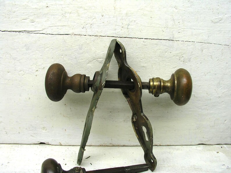 May include: A set of antique brass door hardware featuring two round knobs connected by a square spindle, accompanied by two elongated metal backplates with keyhole cutouts. The metal shows a weathered patina with visible signs of age and wear.