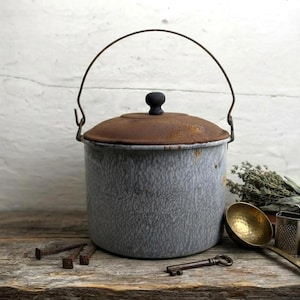 May include: A cylindrical gray enamel pot with a speckled finish, featuring a rusted metal lid with a black knob and a wire bail handle. The pot shows signs of wear and oxidation, with visible rust spots on the lid and along the rim.
