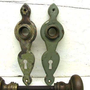 May include: Two vertical sage green metal door plates with keyholes and circular openings for knobs, featuring worn paint and exposed dark metal. Below them, a horizontal metal door spindle connects two round, dark brown metal door knobs.