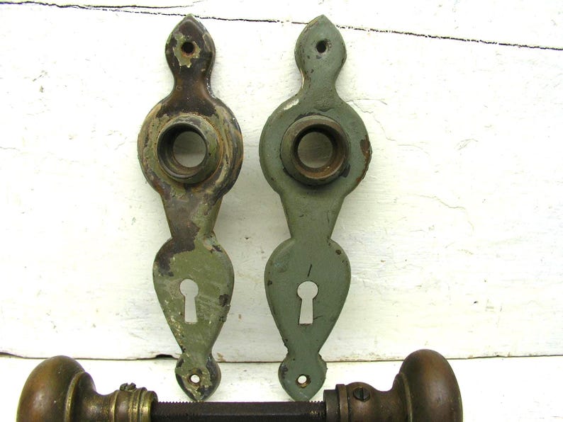May include: Two vertical sage green metal door plates with keyholes and circular openings for knobs, featuring worn paint and exposed dark metal. Below them, a horizontal metal door spindle connects two round, dark brown metal door knobs.