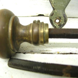 May include: A round, tarnished brass door knob with a smooth, spherical handle and a cylindrical neck. A long, dark, square-profile metal spindle extends from the center of the knob. A separate, matching metal spindle lies horizontally below it.