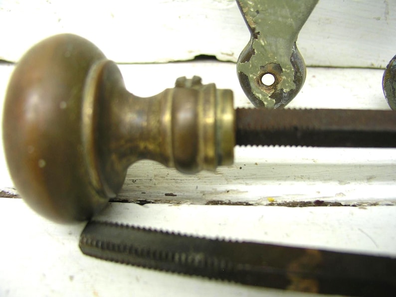 May include: A round, tarnished brass door knob with a smooth, spherical handle and a cylindrical neck. A long, dark, square-profile metal spindle extends from the center of the knob. A separate, matching metal spindle lies horizontally below it.