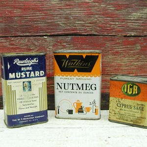May include: Three vintage spice tins: Rawleigh's Pure Mustard, Watkins Nutmeg, and IGA Cyprus Sage. The mustard tin is blue and white, the nutmeg tin is orange and white, and the sage tin is orange and beige. The tins are on a white surface.
