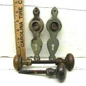 May include: Two vintage door plates with a sage green finish and keyholes, paired with two dark brown metal door knobs on spindles. A wooden ruler is positioned vertically next to the plates, showing a measurement of approximately 7 inches.