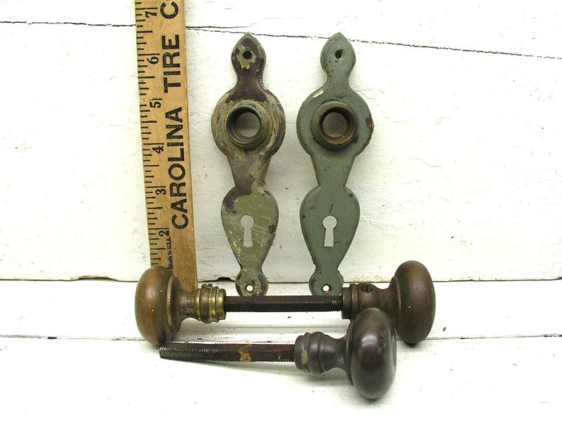 May include: Two vintage door plates with a sage green finish and keyholes, paired with two dark brown metal door knobs on spindles. A wooden ruler is positioned vertically next to the plates, showing a measurement of approximately 7 inches.