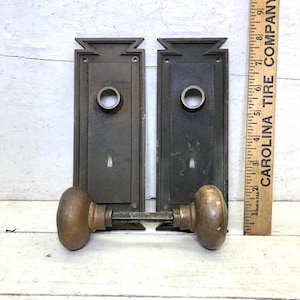 May include: Two antique bronze door handle sets. Each set includes a rectangular backplate with a keyhole and a circular knob. The knobs are connected by a metal rod. A wooden ruler with text is in the background.