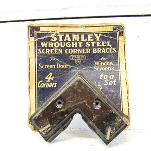 May include: Vintage Stanley wrought steel screen corner braces. The dark metal corner brace is in front of a vintage cardboard package. The package has text that reads "STANLEY WROUGHT STEEL SCREEN CORNER BRACES" and "for Screen Doors".