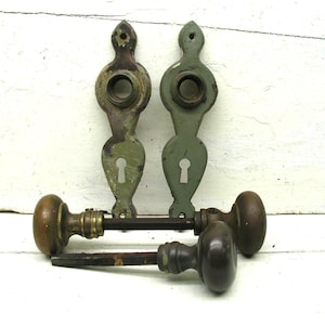May include: Two vertical sage green metal door plates with keyholes and circular openings, paired with two brown round door knobs attached to metal spindles. The hardware shows signs of wear and aged patina, presented as a set of door components.