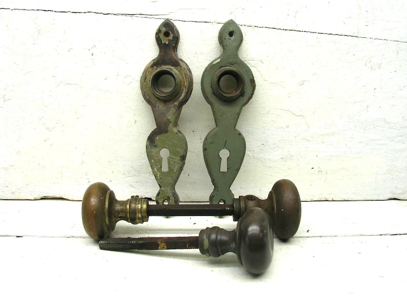 May include: Two vertical sage green metal door plates with keyholes and circular openings, paired with two brown round door knobs attached to metal spindles. The hardware shows signs of wear and aged patina, presented as a set of door components.