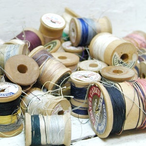 May include: A collection of vintage wooden spools of thread in various colors, including black, white, red, blue, and pink. The spools are labeled with the brand "COATS & CLARK'S" and "COATS" in white lettering on a blue background.