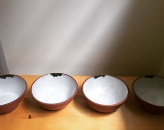 May include: Four terracotta bowls with white glaze and a green rim. The bowls are arranged in a row on a wooden surface.