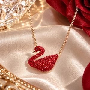 May include: A gold-toned chain necklace featuring a swan-shaped pendant encrusted with small, vibrant red crystals. The swan has a smooth, metallic beak and is suspended from a delicate link chain, creating a refined and detailed jewelry piece.