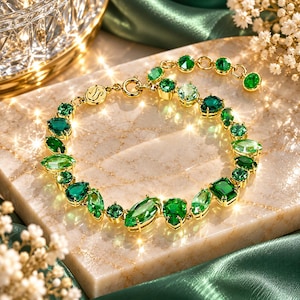 Op de afbeelding: Een goudkleurige armband met een reeks gefacetteerde groene edelstenen in verschillende vormen, waaronder peer-, marquise- en ronde slijpvormen. De ketting bevat een ronde sluiting en een kleine ronde bedel met een gegraveerd symbool.