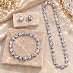 Puede incluir: Un conjunto de joyería compuesto por un collar, una pulsera y un par de pendientes de botón. Cada pieza presenta grupos circulares de piedras claras y brillantes engastadas en metal plateado, con eslabones rectangulares de corte baguette.