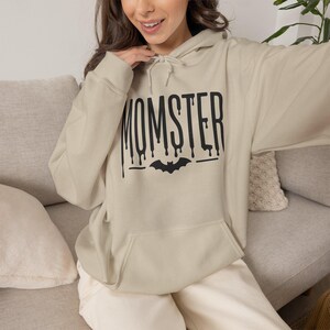 Peut inclure: Un sweat à capuche beige avec une impression graphique noire sur la poitrine. Le texte MOMSTER est écrit dans une police dégoulinante, placé au-dessus d'une petite silhouette de chauve-souris noire. Le vêtement possède une poche kangourou.