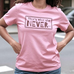 Może przedstawiać: Jasnoróżowy t-shirt z krótkim rękawem z prostokątną brązową ramką zawierającą napis "That's what she NEVER said" pogrubioną, ciemnobrązową czcionką. Koszulka ma okrągły dekolt i standardowy krój z wyraźnym, czytelnym nadrukiem tekstowym.