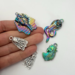 May include: A collection of four metal charms featuring ancient Egyptian motifs. One charm is a rainbow-coloured pharaoh mask, one is a rainbow-coloured cobra, one is a rainbow-coloured profile of a queen, and one is a silver-toned pharaoh mask.