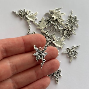 May include: Silver-toned fairy charms with detailed wings and flowing dresses. These charms are ideal for jewelry making or craft projects. The charms are small, with a delicate design, and are shown scattered on a white surface.