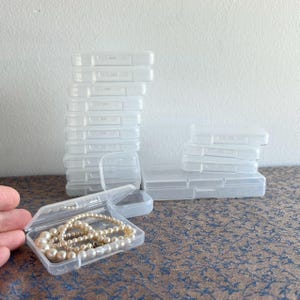 May include: A collection of small, clear plastic hinged boxes. One box is open, displaying a pearl necklace. The boxes are stacked in various arrangements, suggesting storage or organizational use. The background is a neutral white wall.