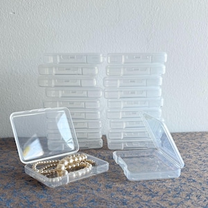 May include: Clear plastic rectangular storage boxes, some stacked, with hinged lids. One box is open, revealing a pearl necklace. The boxes are ideal for organizing small items like jewelry, beads, or craft supplies.