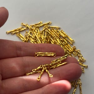 May include: Gold-colored jewelry findings, including textured bar connectors and dangling components with small loops. These components are likely used in jewelry making for necklaces, bracelets, and earrings. The findings have a hammered or organic texture.