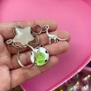 以下が含まれることがあります： 星、恐竜、緑色のアクセントが付いたサッカーボールを特徴とするキーホルダーのコレクション。キーホルダーはシルバーで、シルバーのキーリングに取り付けられています。背景はピンクです。