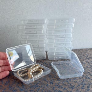 May include: Clear plastic square jewelry boxes, some open and some closed, stacked in two piles. One box contains a pearl necklace. The boxes are on a patterned surface.