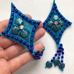 May include: Two blue star-shaped fabric embellishments with blue beads and metallic seashells. Each embellishment has a dangling fringe of blue beads.