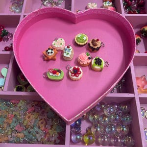 May include: A selection of miniature dessert charms, including cakes and tarts, displayed in a pink heart-shaped box. The charms feature various toppings and colours, such as strawberries, blueberries, and chocolate. The box is set within a larger organiser.