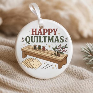 May include: A round white ceramic ornament with a white ribbon hanger. The design features the text HAPPY QUILTMAS in red and green, a wooden sewing bench with three spools of thread, a sprig of holly, four fabric squares, and two sewing needles.