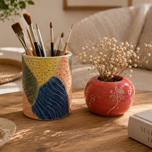 Peut inclure: Un porte-crayon cylindrique en céramique avec un motif géométrique texturé jaune, bleu, vert et pêche, contenant divers pinceaux, à côté d'un petit vase rond en céramique rouge rempli de fleurs blanches séchées.